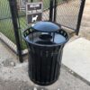 A black metal trash can with a dome lid stands in front of a chain-link fence and a sign that reads "Welcome to the Dog Park" with a white dog silhouette. Gravel and concrete are visible on the ground.