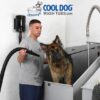 A man uses a blow dryer to dry a wet German Shepherd in a stainless steel dog wash tub. The "Cool Dog Wash Tubs" logo with a cartoon dog and sheep is displayed at the top of the image.