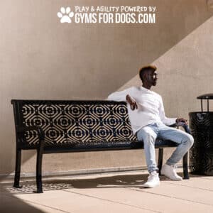 A man in a white shirt and jeans sits on The Luxury Park Bench - Thermoplastic Coated Steel - 6ft near a wall. Above him, text reads, "Play & Agility Powered by Gyms for Dogs.com" with a paw print icon.