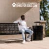 A man in a white shirt and light jeans sits on a patterned outdoor bench near a black trash can, with sunlight casting shadows. "Play & Agility Powered by Gyms For Dogs.com" is displayed above him.
