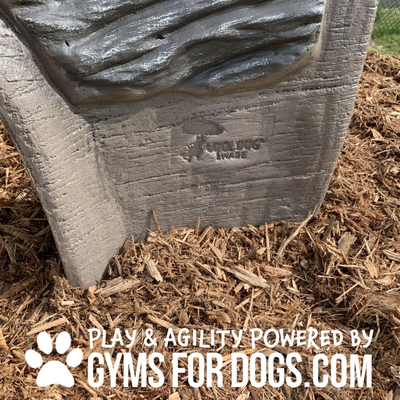 Close-up of a textured, gray structure set in mulch, with the embossed text "COOL DOG MADE" and the words “Play & Agility Powered by GymsForDogs.com” in bold white letters at the bottom.