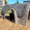 A black dog lies in the shade inside a faux-log dog house at a park, surrounded by wood chips and modern apartment buildings in the background.