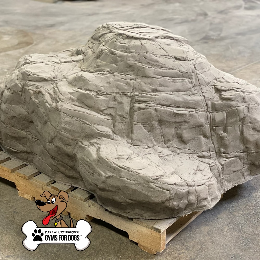 A large artificial rock structure sits on a wooden pallet indoors. The texture mimics real stone. In the corner, there is a “Gyms For Dogs” logo with a cartoon dog and a bone.