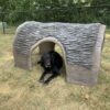A black dog lies under a large, hollow, artificial log in a grassy, fenced yard with trees in the background.
