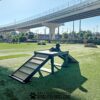 A black dog rests atop the Bridge Climb (Pro), a wooden agility platform, in a grassy dog park with city buildings and an overpass in the background as sunlight casts long shadows across the scene.