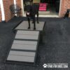 A black dog stands on a Bridge Climb (S/M) 18" Tall agility platform with ramp outdoors on pavement; another black dog sits nearby. The "Gyms for Dogs" logo is displayed in the bottom right corner.