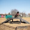 A black dog with a red bandana sits on the Bridge Climb 29" Tall (L/X) at the dog park, surrounded by ramps, a green tunnel, and chairs, all beneath a clear sky.