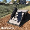 A black dog runs down a Bridge Climb 29" Tall (L/X) agility ramp at an outdoor dog park with wood chips and fencing. The image features the “Play & Agility Powered by Gyms For Dogs.com” logo and text in the corner.