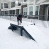 A black dog in a red harness stands on a snowy Bridge Climb 29" Tall (L/X) ramp at a Dog Park near an apartment building, while snow falls and partially covers the ramp.