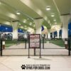 A fenced dog park with green turf sits under a large overpass at dusk, lit by overhead lights. Near the entrance stands the Professional Rules & Wellness Sign - Surface Mount - Vertical Sign. Text at bottom: "Play & Agility Powered by GymsForDogs.com.