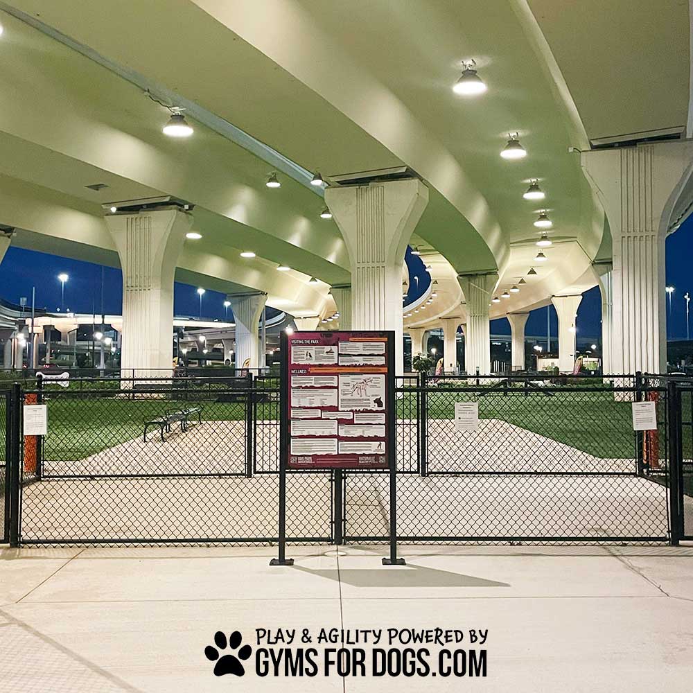 A fenced dog park with green turf sits under a large overpass at dusk, lit by overhead lights. Near the entrance stands the Professional Rules & Wellness Sign - Surface Mount - Vertical Sign. Text at bottom: "Play & Agility Powered by GymsForDogs.com.