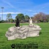 Two dogs, one black and one white with brown spots, sit on large Interactive Pack Boulders - Interactive Play Series in a grassy park with trees and sports fields. Text at the bottom reads, "Play & Agility Powered by Gyms for Dogs.