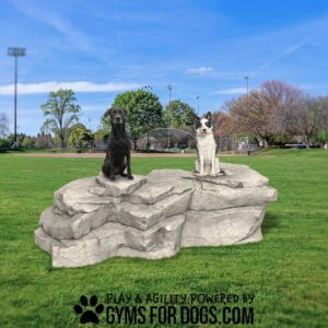 Two dogs, one black and one white with brown spots, sit on large Interactive Pack Boulders - Interactive Play Series in a grassy park with trees and sports fields. Text at the bottom reads, "Play & Agility Powered by Gyms for Dogs.