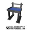 A black and blue elevated dog bench with back support, branded with "GYMS FOR DOGS." Below it are the words "PLAY & AGILITY POWERED BY GYMSFORDOGS.COM" with a black paw print graphic.