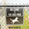 A 15"x15" Dog Park Welcome Sign is mounted on a metal fence, featuring black and white text, a running dog graphic, "Play & Agility Powered by GymsForDogs.com," with grass and a paw print sculpture visible behind it.
