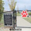 A black chain-link fence gate to a dog park displays a sign with dog park rules and a large red paw print sign. Trees and a walking path are visible inside the park.