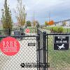 A black chain-link fence with signs reading "Bark Park at The Mark" and "Welcome to the Dog Park" marks the entrance to a dog park on a cloudy day, with grass, trees, and houses visible in the background.