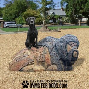 A black dog sits atop a large, gorilla-shaped Ranger - Interactive Play Series structure in a park, surrounded by grass, trees, and houses. Text at bottom: "Play & Agility Powered by GymsForDogs.com.