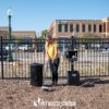 A woman in a yellow shirt uses The Ultimate Park Caddy System—including Waste Caddy & Storage Caddy with Scooper—to clean pet waste at a dog park by a black waste station and trash bin, with buildings and a fence in the background.