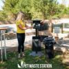 A woman in a yellow shirt stands at The Pet Waste Station - Surface Mount in a fenced dog park, holding a roll of waste bags. Nearby are a trash can and a sign that reads "PET WASTE STATION.
