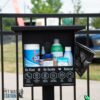 A black pet waste station attached to a fence, stocked with cleaning supplies, disinfectant wipes, sprays, treats, and poop bags. Signs below read "Be Kind," "Be Gentle," and "Natural." Outdoor scene in background.