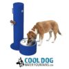 A brown and white dog drinks from a blue Cool Dog Water Fountain - SST - Dog Basin & Hose Bib, featuring a bowl and spout. Below the fountain is the "Cool Dog Water Fountains" logo with a dog illustration.