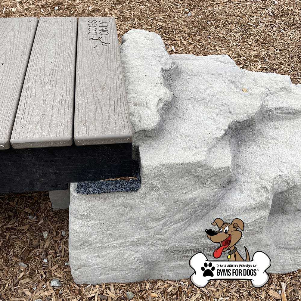 A playground feature with a wooden bench connected to an artificial rock structure, surrounded by wood chips. The "Gyms for Dogs" logo with a cartoon dog appears in the lower right corner.
