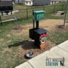 A pet waste station in a park with a green dispenser for waste bags, a decorated black bin with a flower pot design, a metal dog bowl on the ground, and a fence and houses in the background.