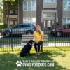 A woman in a yellow shirt sits on a bench in a grassy area, smiling and petting a black dog. Behind them is a black fence, parked cars, and an apartment building. Text reads: "PLAY & AGILITY POWERED BY GYMS FOR DOGS.COM.