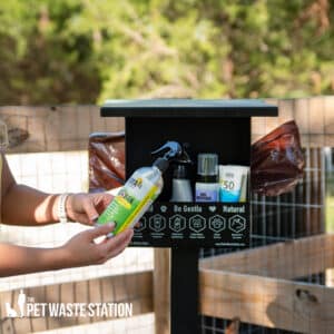 A person holds The Wellness Depot - First Aid, Wound Care Convenient Storage Container near a pet waste station with bags, sanitizer, and wipes outdoors by a wooden fence and trees. The Pet Waste Station logo appears at the bottom left.