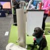 A black dog wearing a teal harness sits beside a modern water fountain designed for humans and pets at an indoor event with people and booths in the background.