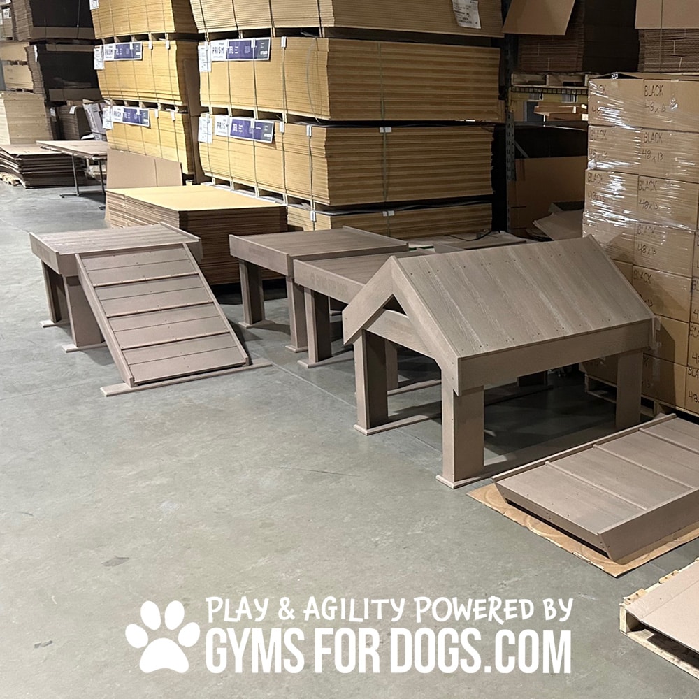 A warehouse with stacks of cardboard boxes contains several brown wooden dog agility ramps and platforms for a Dog Park. The text reads: "PLAY & AGILITY POWERED BY GYMSFORDOGS.COM" with a paw print.