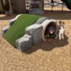 Two dogs sit next to a playground structure resembling a small stone tunnel with a grassy hill on top, surrounded by wood chips.