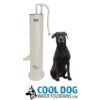 A black dog sits beside a tall, silver and white Doggie Demand Fresh Flow Portable Water Fountain Spout & Pet Hose (SLV) at a dog park. Below is the Cool Dog Water Fountains logo with a dog illustration and blue text.