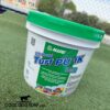 A white tub of Mapei Ultrabond Turf PU 1K adhesive with a green lid sits on artificial grass. The label features product details and caution signs. A fence and buildings are visible in the blurred background.